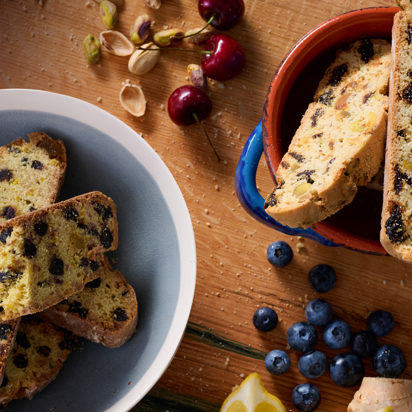 Two boxes of biscotti, one labeled Blueberry + Pistachio and the other Cherry + Pistachio, arranged on a wooden surface with accompaniments such as cherries, blueberries, pistachio nuts, lemon slices, and ginger.